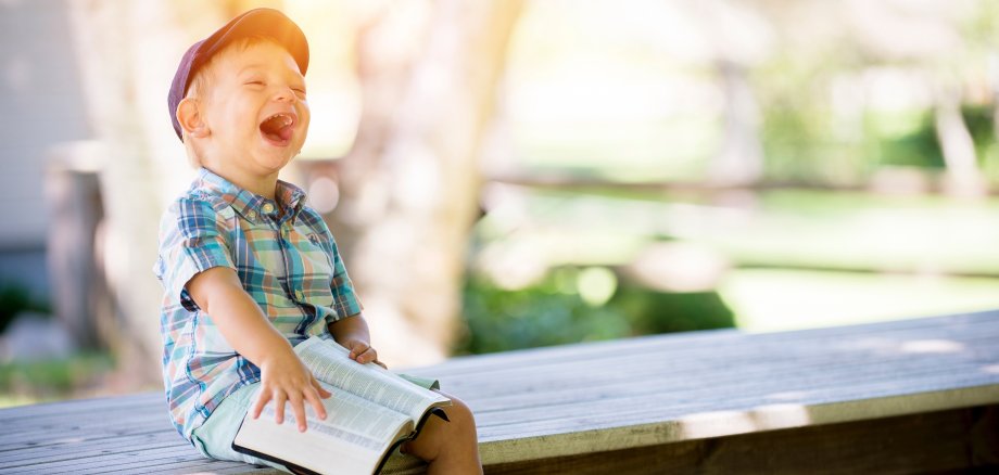 Ein kleiner Junge sitzt auf einer Bank mit einem Buch auf dem Schoß und lacht