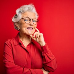 Senior beautiful grey-haired woman wearing casual shirt and glasses over red background looking confident at the camera with smile with crossed arms and hand raised on chin. Thinking positive. Senior beautiful grey-haired woman wearing casual shirt and glasses over red background looking confident at the camera with smile with crossed arms and hand raised on chin. Thinking positive.